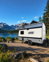  surrounded by forest with a picnic table and fire pit on a gravel site.|This Coleman travel trailer RV rental