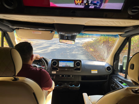 An RV rental interior: a man drives on a leaf-covered road, a football game visible on the overhead TV, merging travel & entertainment.