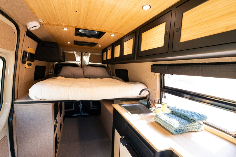 RV rental interior with an elevated bed, kitchenette with sink, overhead cabinets, and warm wood accents throughout. — photo 7
