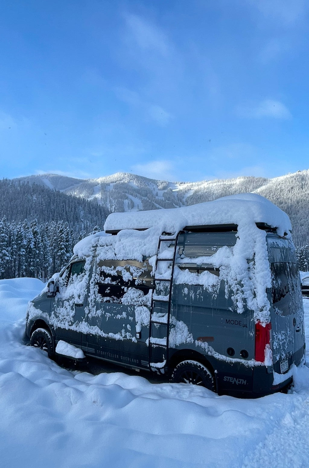 A gray Storyteller Overland van, suitable for an rv rental adventure, is heavily snowed in with deep drifts & snowy mountains behind it.