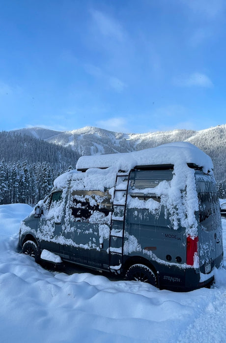 A gray Storyteller Overland van, suitable for an rv rental adventure, is heavily snowed in with deep drifts & snowy mountains behind it.