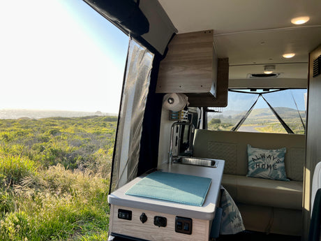 RV rental interior: A compact kitchen and seating area with a screen door open to a sunny coastal view with green grass and ocean.