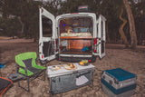 This RV rental shows an open camper van with a cozy interior, an outdoor kitchen setup with a Dometic cooler and snacks in a natural setting.
