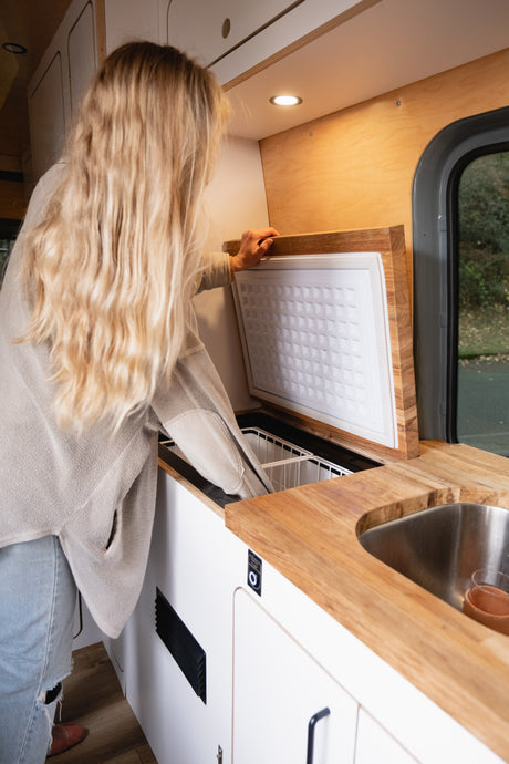 Inside an rv rental: A blonde woman opens a top-loading cooler in a stylish camper van kitchen setup.