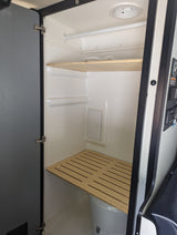 RV rental compact wet bath: toilet under slatted wooden bench, upper shelf, and shower rod. Practical interior for travel.