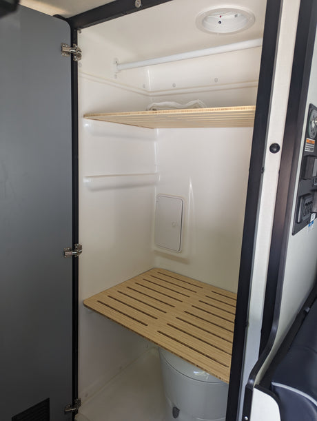 RV rental compact wet bath: toilet under slatted wooden bench, upper shelf, and shower rod. Practical interior for travel.
