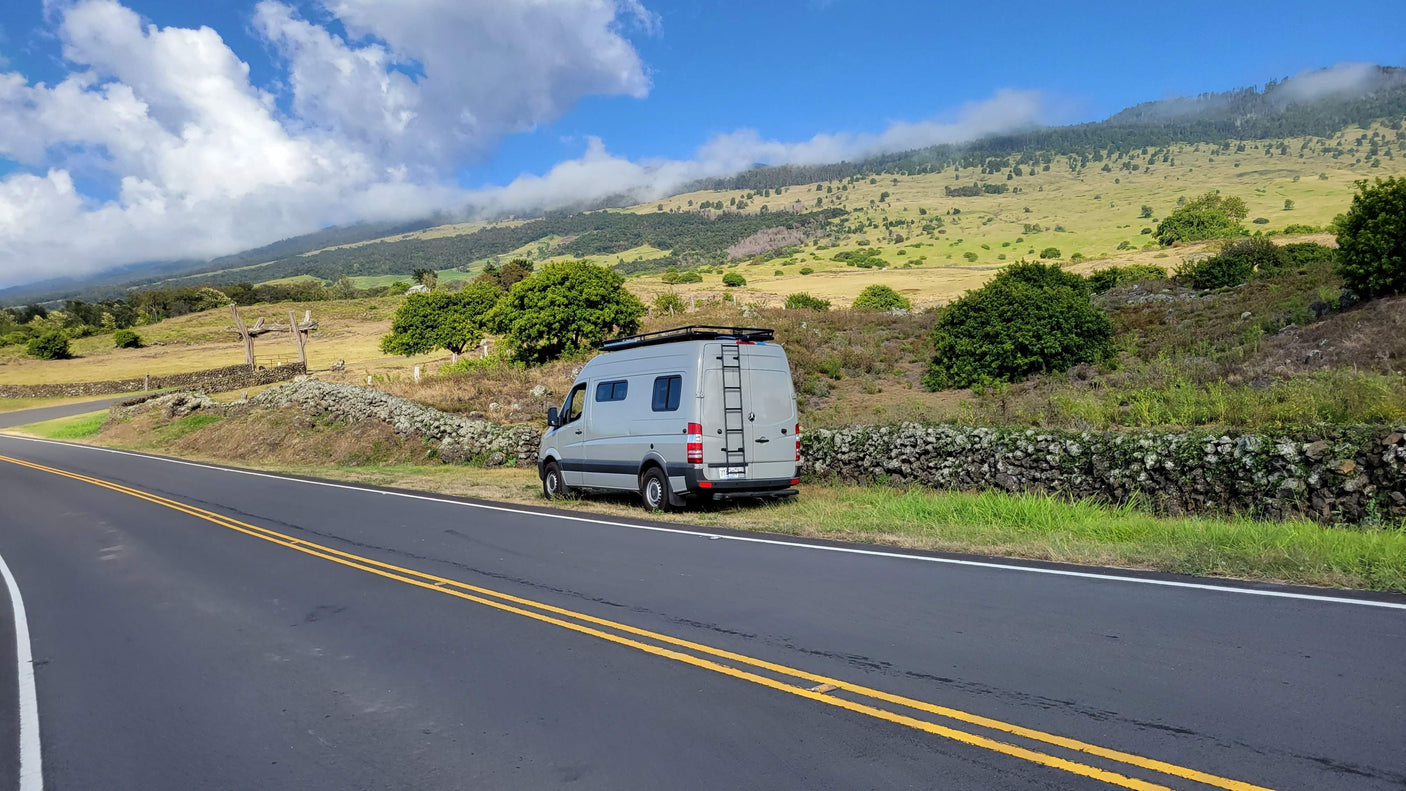 Grey RV rental on scenic roadside. Green mountains, clouds, and stone wall mark a picturesque landscape.