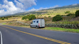 Grey RV rental on scenic roadside. Green mountains, clouds, and stone wall mark a picturesque landscape.