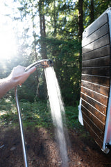 Hand holding chrome shower head spraying water in a sun-dappled forest, part of an RV rental outdoor setup.