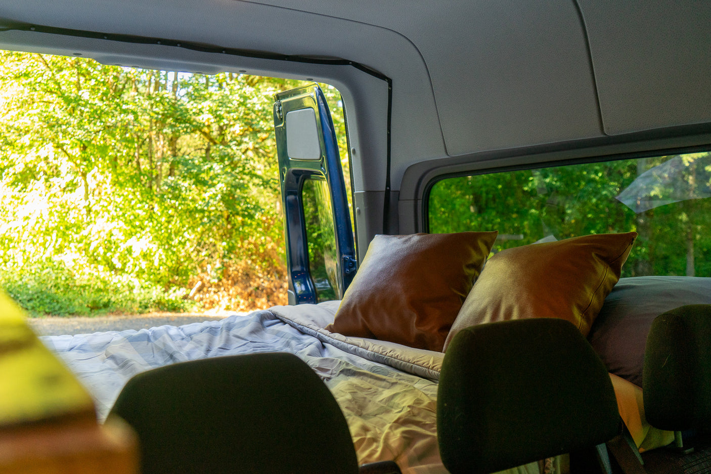 Inside an rv rental: a cozy bed with brown pillows, seen from front seats. Open side door reveals lush forest. Great for travel!