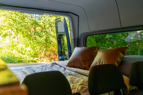 Inside an rv rental: a cozy bed with brown pillows, seen from front seats. Open side door reveals lush forest. Great for travel!