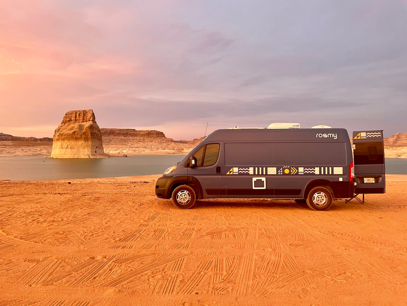 An RV rental van, "Roamy," parked on red sand by a lake with desert rock formations at sunset. Experience nature!