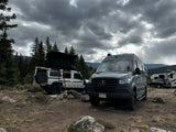 A white SUV with a rooftop tent & a grey camper van (likely an RV rental) parked at a rugged mountain campsite under cloudy skies.