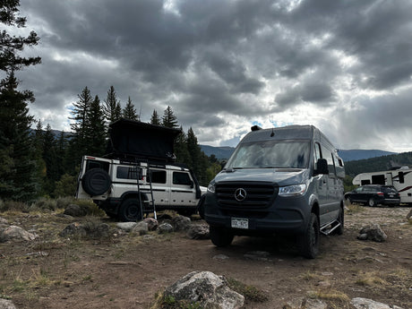 A white SUV with a rooftop tent & a grey camper van (likely an RV rental) parked at a rugged mountain campsite under cloudy skies.