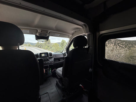 Inside an RV rental, a scenic road trip view through the windshield. Dashboard, front seats, and winding mountain road with trees visible.