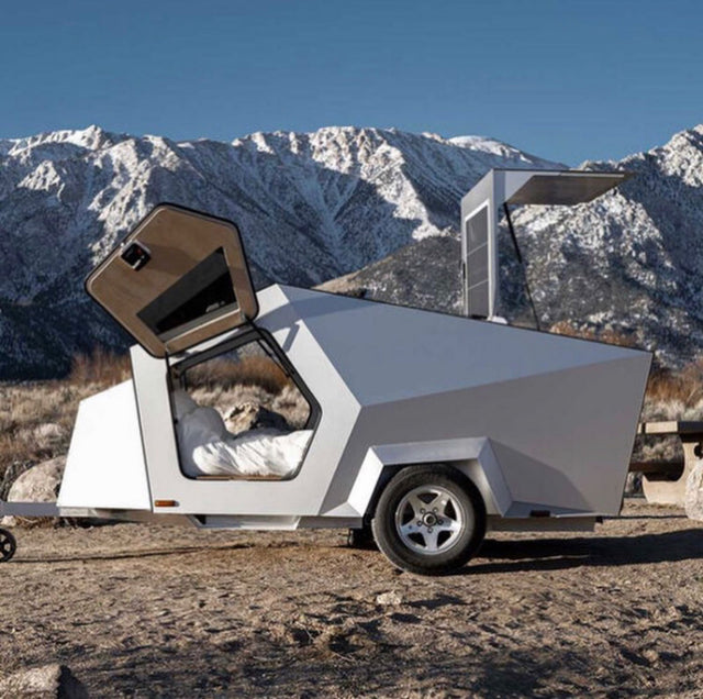 A futuristic, angular travel trailer, perfect for an RV rental, is parked among snow-capped mountains. A bed is visible inside.