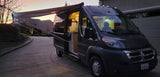 This dark grey camper van, perfect for rv rental adventures, features an extended awning & cozy illuminated interior at sunset.