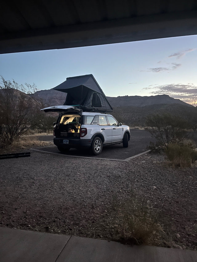 White SUV with rooftop tent deployed, open hatch revealing gear, in a desert campsite at twilight. Consider this RV rental.