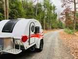 White teardrop RV rental on a gravel road through a dense forest. Perfect for off-grid camping adventures.
