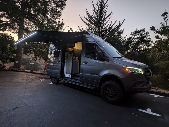 Experience adventure with this gray Mercedes Sprinter rv rental van, featuring an illuminated awning & inviting interior at dusk.