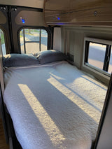 Inside a comfy rv rental: a sunlit bed with grey pillows & white blanket, ready for rest.
