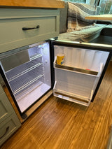 Open, compact fridge in a clean rv rental, showing shelves and integrated green cabinetry, next to a cozy seating area.