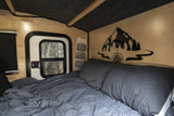 Cozy RV rental interior: comfy bed, window with tree view, and scenic mountain decal, ideal for camping and travel.