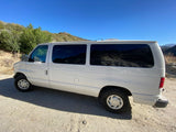 White van, an RV rental, parked on a sunny dirt road amidst green mountains under a clear blue sky.