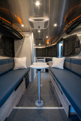 Inside a modern RV rental: a compact dinette with blue benches, central table, and kitchenette. Bright lights illuminate the sleek interior.