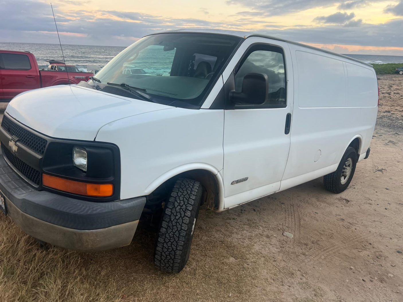White panel van with all-terrain tires on a sandy beach by the ocean. Ideal for an RV rental or beach camping trip.|RV rental interior: van bathed in red LED string lights on ceiling & walls