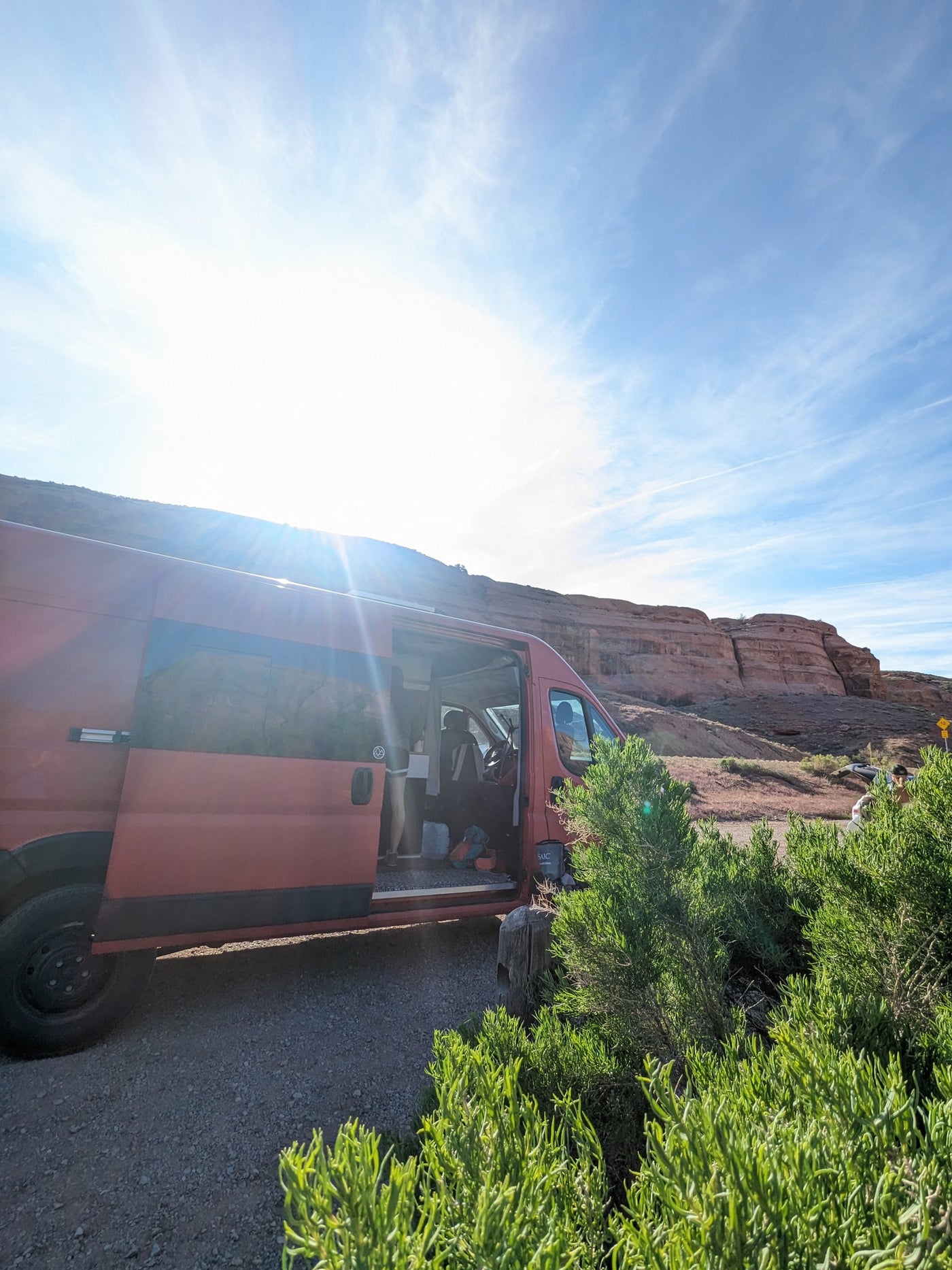 Red camper van parked with open door by red rocky cliffs on a sunny day. Your adventure starts with an RV rental!|This red Ram ProMaster camper van