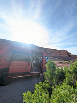 Red camper van parked with open door by red rocky cliffs on a sunny day. Your adventure starts with an RV rental!