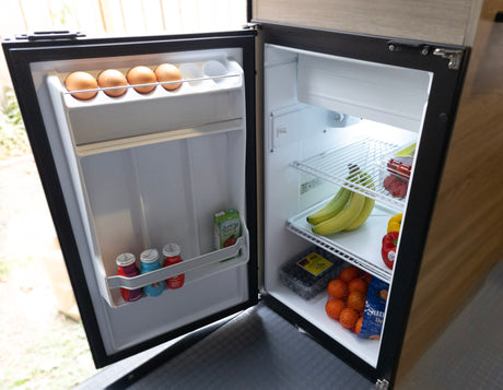 An open RV rental fridge stocked with fresh eggs, fruit (bananas, berries, tangerines), vegetables, and assorted drinks.
