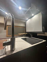Modern RV rental interior: kitchen with sleek faucet, induction cooktop, and large projection screen for entertainment.
