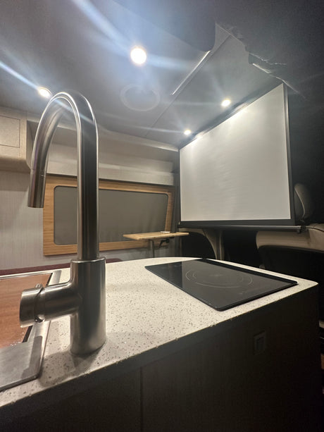 Modern RV rental interior: kitchen with sleek faucet, induction cooktop, and large projection screen for entertainment.