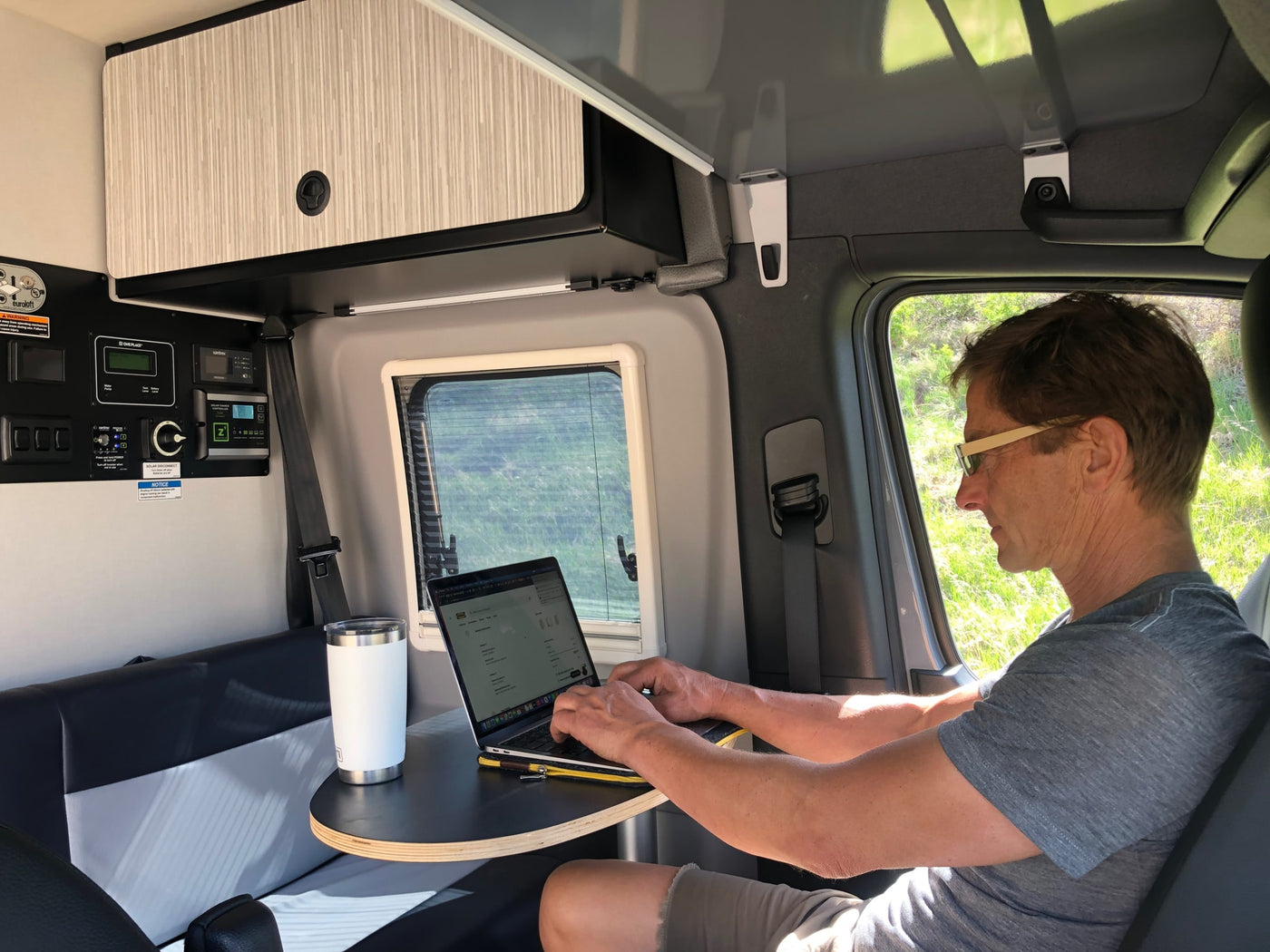 Enjoy remote work in an RV rental! A man uses his laptop at a table inside a camper van, surrounded by controls & nature views. — photo 3