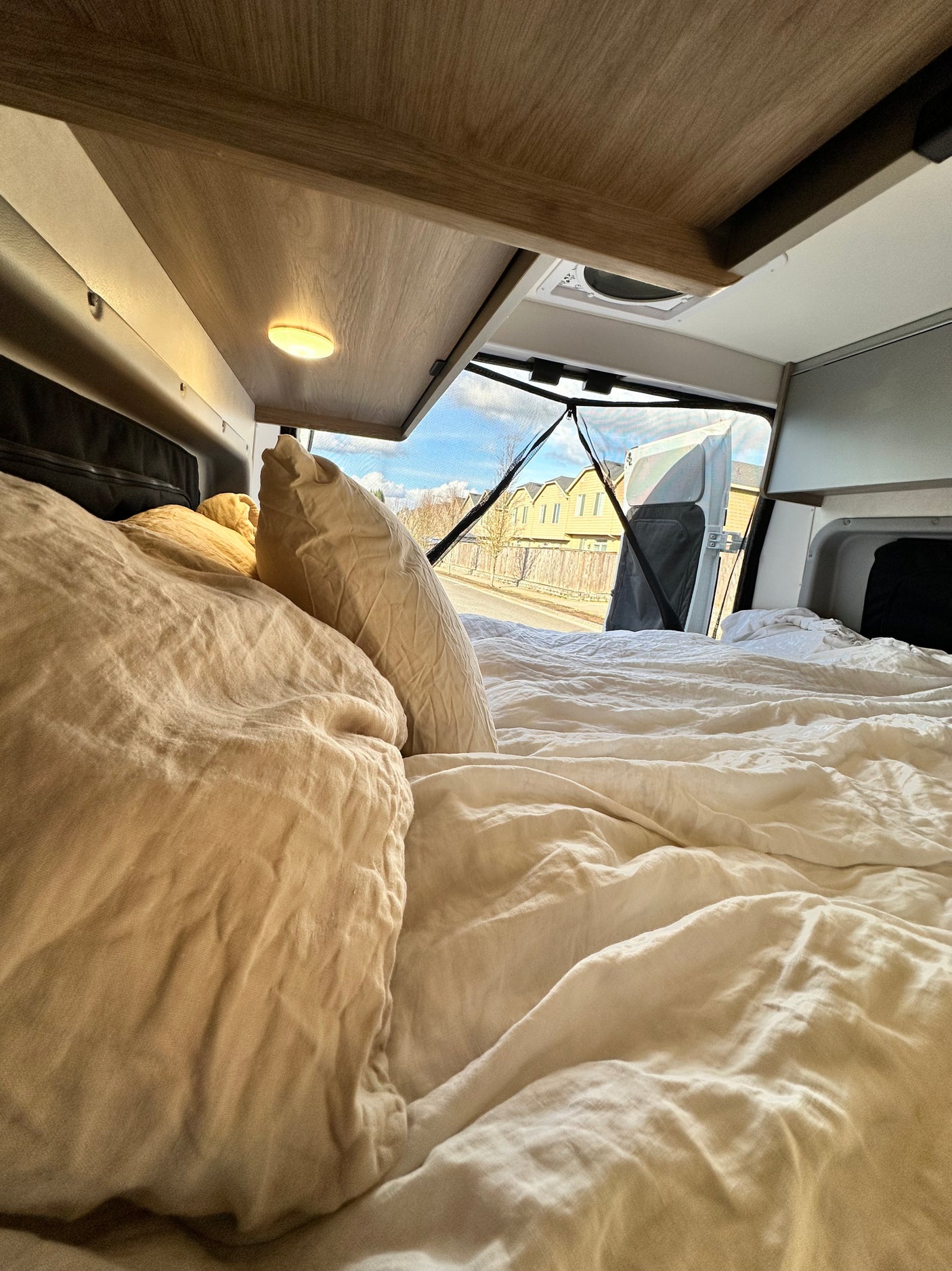 Cozy RV rental interior with a comfy bed, looking out the open rear doors at a street and houses.