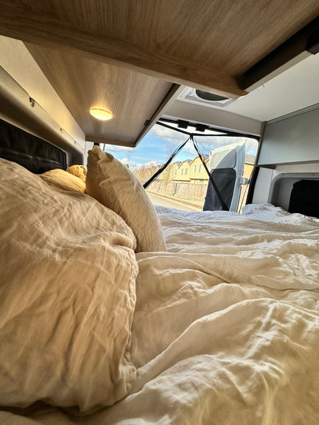 Cozy RV rental interior with a comfy bed, looking out the open rear doors at a street and houses.