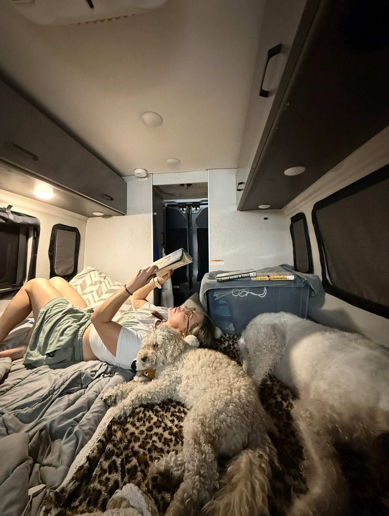 Inside an RV rental: a woman relaxes reading a book, accompanied by two fluffy dogs on a cozy leopard blanket.