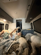 Inside an RV rental: a woman relaxes reading a book, accompanied by two fluffy dogs on a cozy leopard blanket.