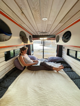 Working remotely from a cozy RV rental: A woman uses her laptop on a bed inside a camper van, overlooking a scenic landscape.