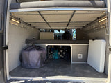 Inside an RV rental, the rear cargo area features an elevated bed, ample storage, side lights, a large black bag, and a white folding table.