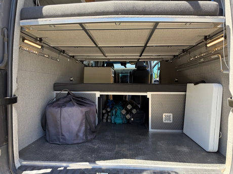 Inside an RV rental, the rear cargo area features an elevated bed, ample storage, side lights, a large black bag, and a white folding table.