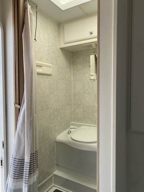 Compact RV rental bathroom with light-tiled walls, a corner toilet, and a grey-striped shower curtain.