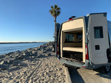 RV rental camper van on a sandy beach by the ocean. Rear doors open, revealing a cozy interior, with a palm tree & ships.