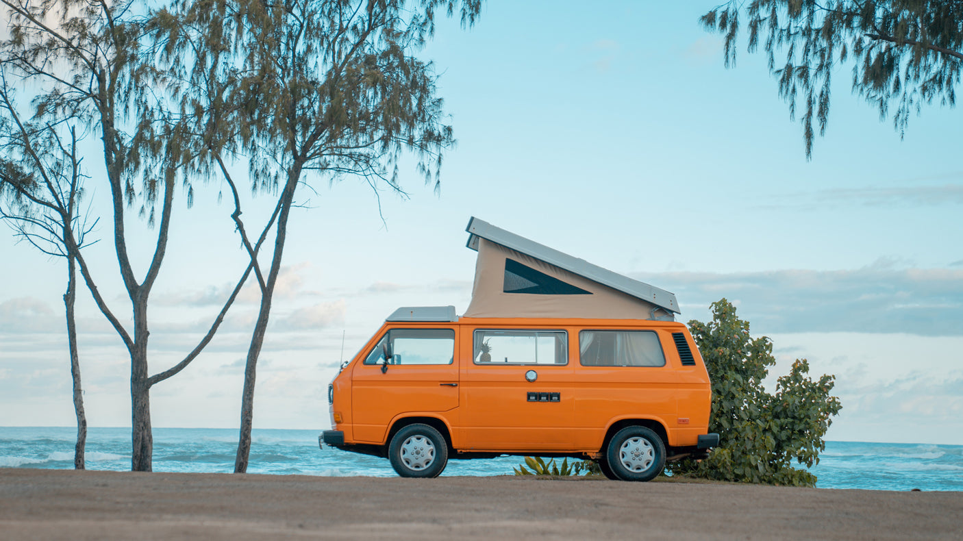 Experience beach life! An orange pop-top camper van for your RV rental, parked near trees by the blue ocean.
