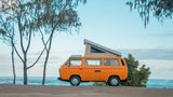 Experience beach life! An orange pop-top camper van for your RV rental, parked near trees by the blue ocean.