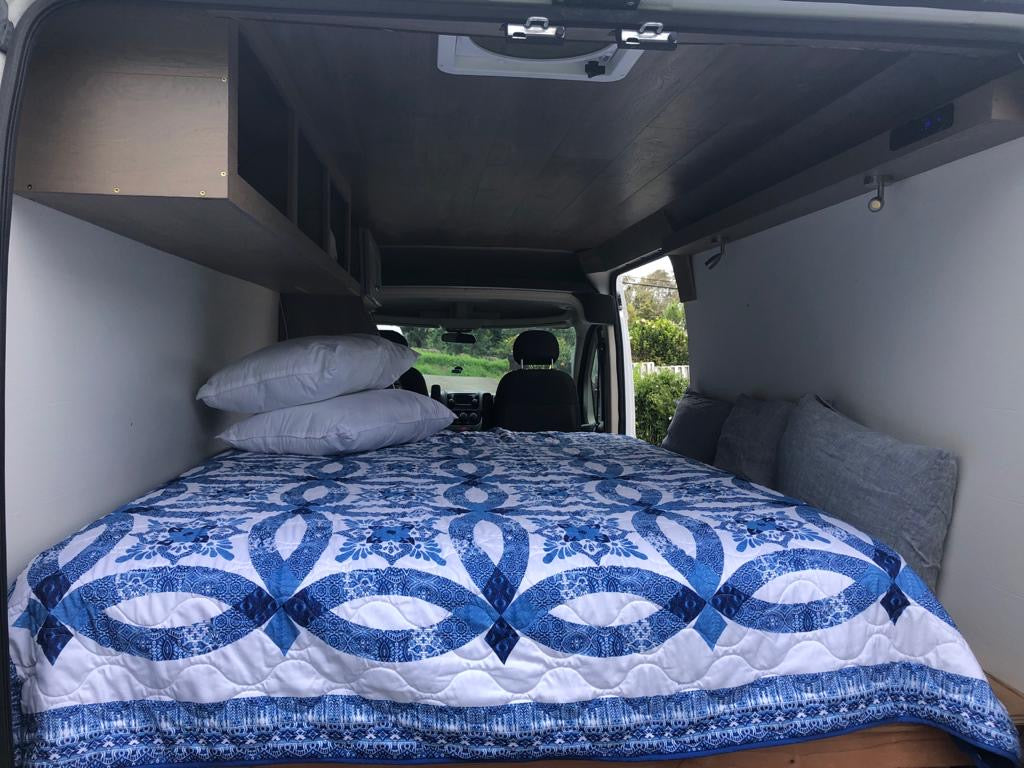 RV rental interior: cozy bed with blue quilt, overhead storage shelves, and reading lights for travel comfort.