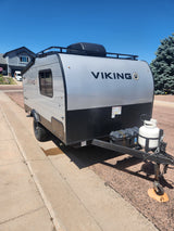 Small Viking travel trailer, possibly an RV rental, with a roof rack, AC unit, and front propane tank, parked on a sunny day.