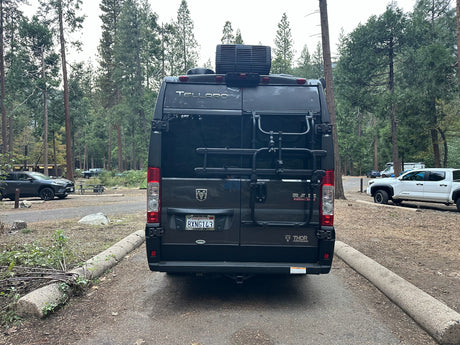 Rear view of a dark gray Thor Tellaro camper van rv rental with a bike rack, parked at a wooded campsite among tall trees.
